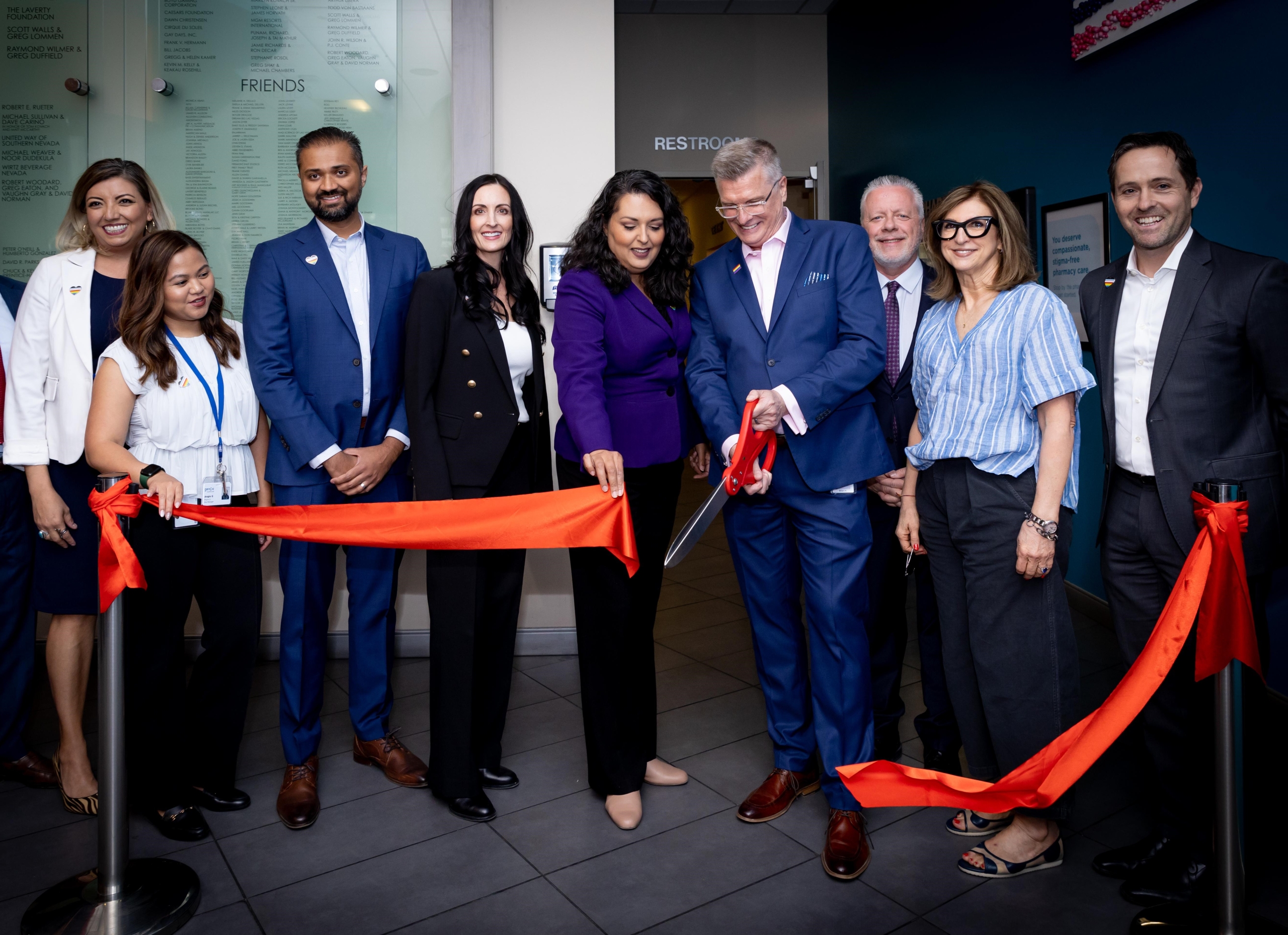 A group of people in business attire stand behind a ceremonious red ribbon. They all adorn heart-shaped rainbow pins on their tops. A man and woman in the center of the group cut the ribbon; the man holds large red scissors while the woman holds the red ribbon.